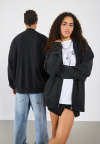 Black zip-up jacket with relaxed fit, paired with a white T-shirt featuring a graphic. Back view of a person in a black top with light denim jeans.
