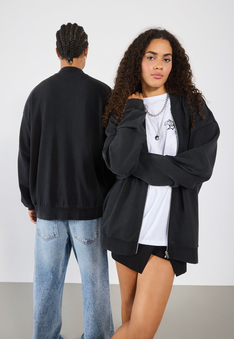 Black zip-up jacket with relaxed fit, paired with a white T-shirt featuring a graphic. Back view of a person in a black top with light denim jeans.
