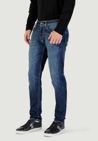 Jeans in denim blu scuro con vestibilità slim, caratterizzati da un classico design a cinque tasche e cuciture gialle a contrasto. Indossati con sneakers nere.