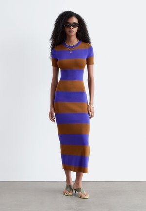 Vrouw met krullend haar met zwarte zonnebril en een aansluitende, korte mouwen, blauw en bruin gestreepte midi-jurk met groene sandalen met hak.