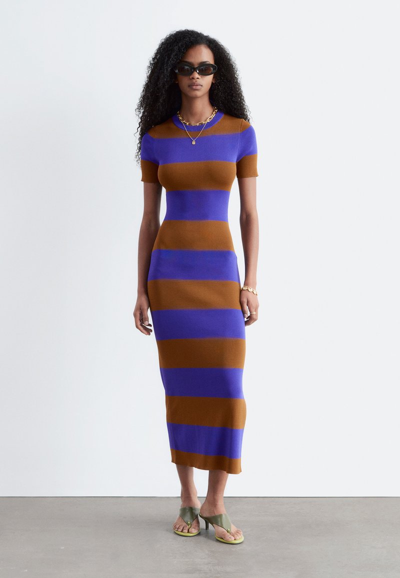 Femme aux cheveux bouclés portant des lunettes de soleil noires et une robe midi ajustée à manches courtes rayée bleu et marron avec des sandales à talons vertes.