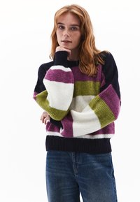 Tjockstickad tröja med breda ränder i lila, grön, vit och marinblå; ribbade muddar och fåll; avslappnad passform. Bärs med blå jeans.