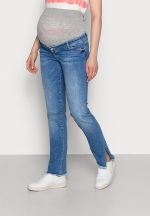 Schwangere Person trägt dehnbare Umstandsjeans mit seitlichen Schlitzen und weiße Sneakers, steht vor einem schlichten Hintergrund.