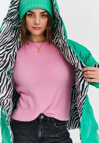 Žena v tealovej čiapke a bunde so zebrovým vzorom vo vnútri, s ružovým sveterom a čiernymi džínami, ktoré drží otvorené, aby ukázala podšívku.