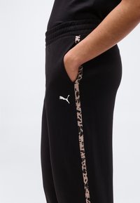 Άτομο που φοράει μαύρη φόρμα Puma με το αριστερό χέρι στην τσέπη και ρίγα με λεοπάρ σχέδιο κατά μήκος της πλαϊνής ραφής.