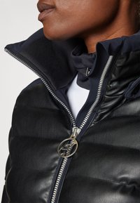 Giacca in pelle nera imbottita con colletto alto, zip argentata e tirazip circolare. Dettagli in tessuto blu navy all'interno.