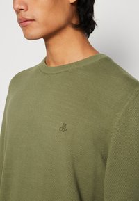 Młoda osoba w oliwkowo-zielonym swetrze o fakturze dzianiny z małym haftowanym logo na piersi, zwrócona w lewo.