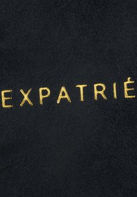 Sort ruskindstof med præget guldt tekst, der staver "EXPATRIÉ." Glat tekstur med mat finish og minimalistisk design.