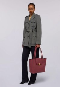 Šedá bunda z vlněné směsi s dvěma náprsními kapsami a pásem v pase, kombinovaná s černými kalhotami a vínově červenou kabelkou na rameno s přezkou.