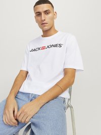 Balta medvilninė marškinėliai su juodu ir raudonu "JACK & JONES" logotipu per krūtinę. Sušvelninta su šviesiai mėlynomis džinsais.