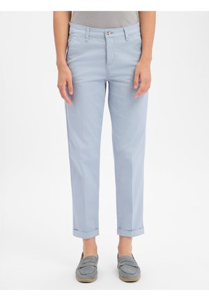 Personne portant un pantalon bleu clair retroussé et des chaussures grises sans lacets, debout devant un fond blanc uni.