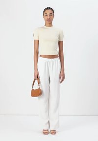 Top beige aderente a maniche corte, pantaloni bianchi larghi e una piccola borsa marrone. Modella in piedi con espressione neutra su uno sfondo bianco.