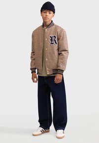 Brown houndstooth bomber jacket with navy accents, a large 'R' emblem, paired with a green top, navy jeans, and white sneakers.