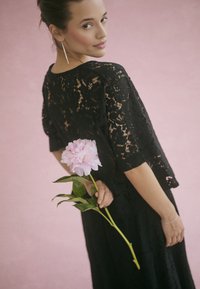 Schwarzes Spitzenkleid mit kurzen Ärmeln, verziert mit Blumenmustern. Das Model hält eine rosa Pfingstrose mit grünen Blättern vor einem pinken Hintergrund.