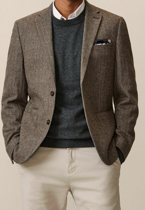 Mann trägt braunen Fischgrätblazer, dunkelgrauen Pullover, weißes Hemd mit Kragen, beige Hose und schwarzes Einstecktuch, Hände in den Taschen.