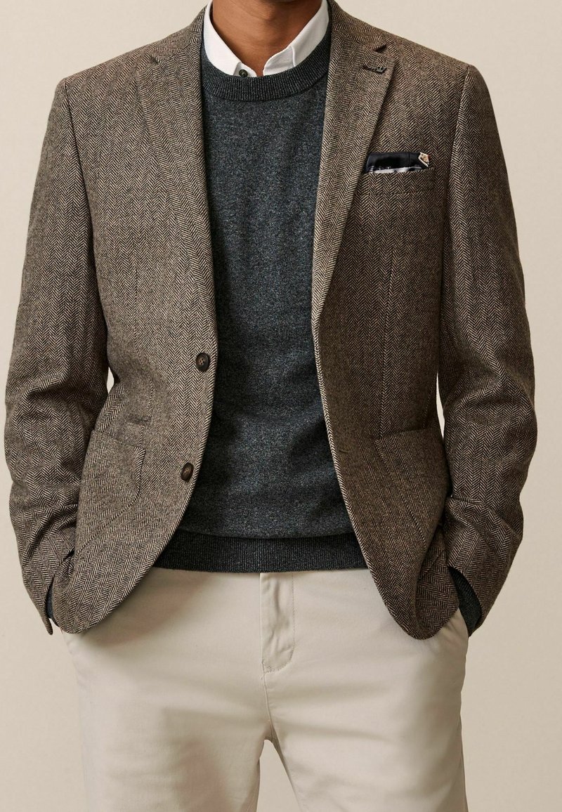 Homme portant un blazer marron en chevron, un pull gris foncé, une chemise blanche col chemise, un pantalon beige et un mouchoir noir en pochette, les mains dans les poches.