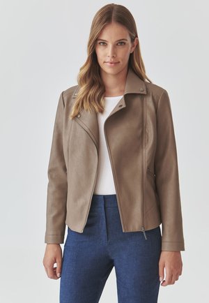 Giacca in finta pelle marrone con un design aderente, zip frontale asimmetrica e bottoni a pressione. Abbinata a una maglietta bianca e pantaloni blu.