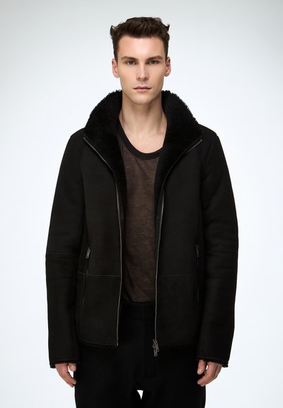 Schwarze Lammfelljacke mit großem Kragen, Frontreißverschluss und zwei Seitentaschen. Getragen über einem dunklen, enganliegenden Hemd.
