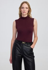 Bordeaux mouwloze coltrui gecombineerd met hoge zwarte nepleer broek, met een gouden gesp en zijzakken.