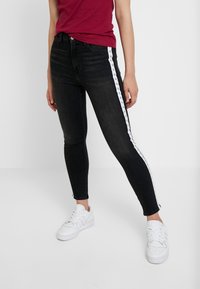 Jeans ajustados negros con rayas laterales blancas que presentan un patrón de logotipos repetido. Los jeans tienen una cintura de altura media y una tela suave y texturizada.
