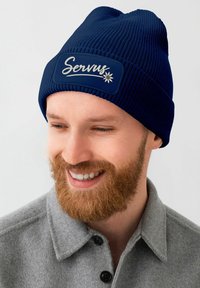 Navy knit beanie with ribbed texture and a rectangular patch displaying "Servus" and a flower design. Features a snug fit.