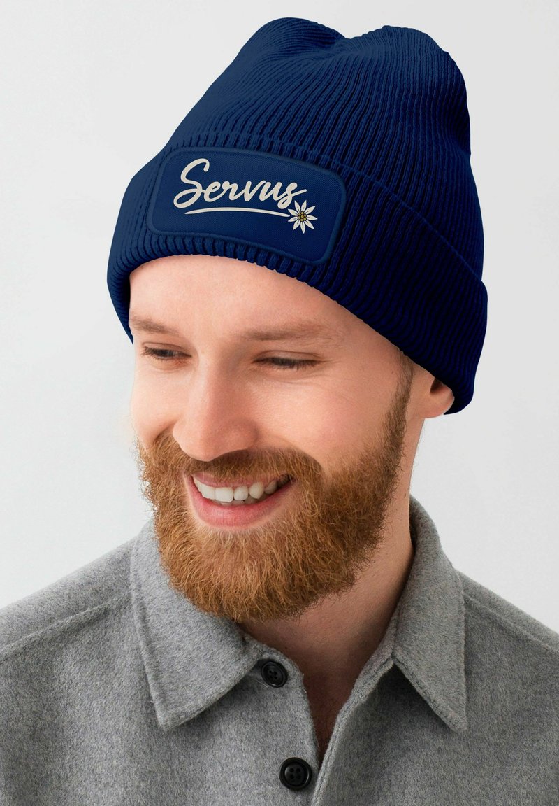 Navy knit beanie with ribbed texture and a rectangular patch displaying "Servus" and a flower design. Features a snug fit.