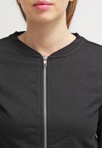 Czarna kurtka zapinana na zamek błyskawiczny z okrągłym dekoltem, wyposażona w srebrny zamek z przodu i gładką, teksturowaną bawełnianą tkaninę.