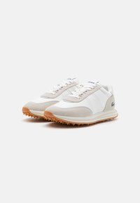 Zapatillas deportivas en blanco y beige que presentan una combinación de materiales lisos y de ante, una suela de goma texturizada y un sutil branding en la lengüeta y el costado.