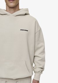 Beige hettegenser med en frontlomme, snorhette og brodert "PECADOR"-logo i svart på brystet; glatt tekstur.