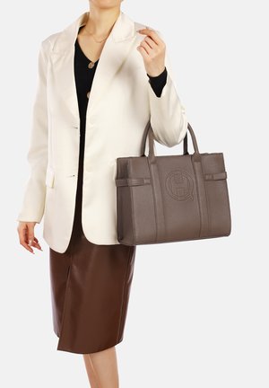 Borsa a tracolla in pelle grigia con superficie testurizzata, caratterizzata da un logo circolare, due manici corti e cinghie decorative. Il modello indossa un blazer bianco e una gonna marrone.