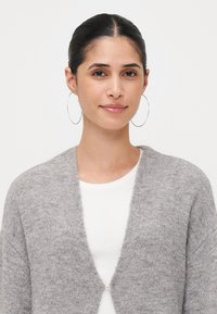Grå strikket cardigan med åpen front, med en enkelt knappelukking. Myk tekstur og ermer av middels lengde, kombinert med en hvit topp.