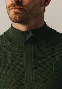 Groene gebreide pullover met een hoge kraag en een halfrits sluiting. Heeft een gestructureerd patroon en een klein logo op de borst.