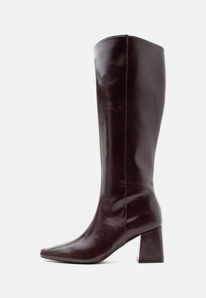 Botte en cuir brun foncé montant jusqu'au genou avec un bout pointu et un talon bloc sur un fond clair uni.