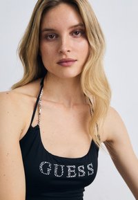 Débardeur noir avec un décolleté arrondi, orné de strass argentés formant le mot "GUESS", avec des bretelles fines embellies de perles.