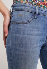 Blå denimjeans med en slät textur, medelhög midja och en subtil sömdesign. Har en liten logotyp på bakfickan.