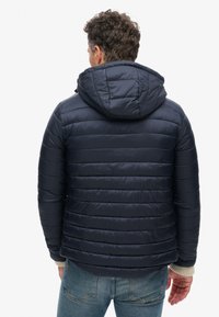 Marineblå polstret jakke med hætte, med vandret syninger, elastiske manchetter og en glat, shiny finish. Bagklap vist.