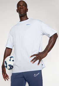 Lyseblå kortærmet t-shirt med "INTER" og Nike-logo i mørkeblå, parret med navy bukser og holder en mønstrede fodbold.