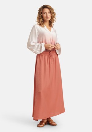 Lange, vloeiende koraalkleurige rok met een tailleband met trekkoord, gecombineerd met een wit-lichtroze ombre blouse met pofmouwen en een V-hals. Platte bruine sandalen.