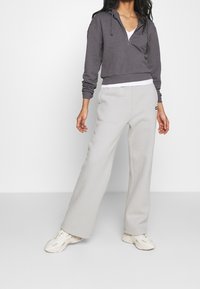 Felpa grigia corta con zip frontale, cordini e una maglietta bianca sotto. Pantaloni da jogging larghi grigio chiaro e sneakers color crema.