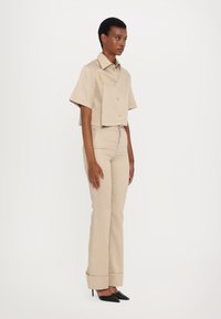 Beige rövidujjú cropped ing, elöl gombos záródással, bézs testre simuló nadrággal és fekete hegyes sarkú cipővel párosítva.