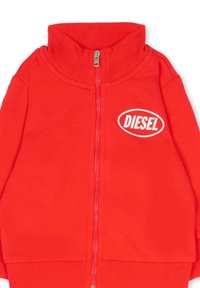 Rode jas met ritssluiting gemaakt van gladde stof, voorzien van een hoge kraag en geribbelde manchetten. Heeft een wit logo aan de linkerkant.