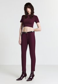 Junge Frau mit welligem dunklem Haar, die ein passendes burgunderfarbenes Crop-Top, schmale Hosen mit einem schwarzen Gürtel und spitze Stiefeletten trägt.