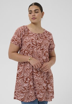 Žena v krátkom rukáve, hnedo-ružovej tunike so vzorom paisley, zlatým náramkom a malými zlatými kruhovými náušnicami na jednoliatom pozadí.