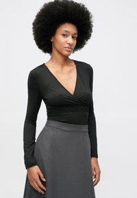 Vrouw met afro-haarstijl, gekleed in een zwarte lange mouwen V-hals top en een hoge taille grijze rok, staande tegen een effen witte achtergrond.