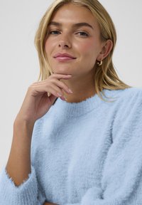 Jasnoniebieski, puszysty sweter z okrągłym dekoltem i długimi rękawami. Miękka tekstura, z lekko falbaniastym wykończeniem dekoltu i mankietów.