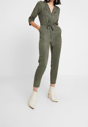 Olivgrüner Jumpsuit mit Reißverschlusstaschen auf der Brust, tailliertem Bund und maßgeschneiderten knöchelangen Hosen, kombiniert mit weißen, hochhackigen Stiefeletten.