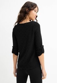 Mujer con cabello ondulado que lleva una blusa negra de manga larga texturizada con puños enrollados, de pie de perfil frente a un fondo blanco.