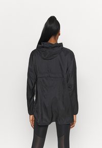 Zwarte lichtgewicht jas met capuchon, voorzien van een gestructureerde stof, lange mouwen en twee zijzakken. Achteraan zichtbaar met een relaxte pasvorm.