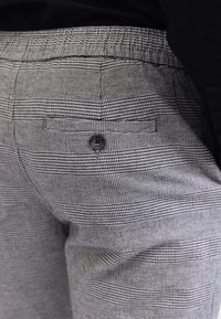 Gros plan sur un pantalon à carreaux gris avec une poche arrière noire boutonnée et une ceinture, porté avec un haut noir.