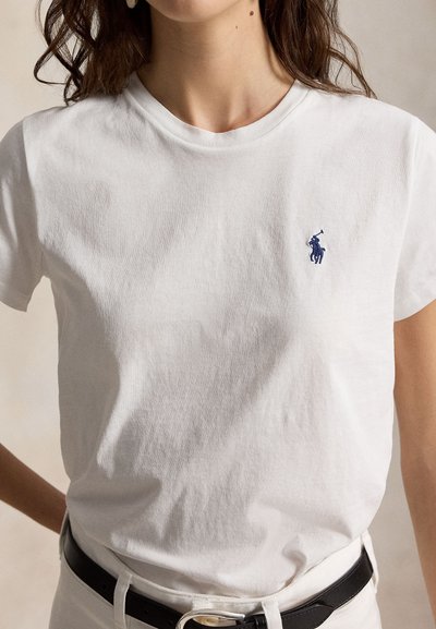 Γυναίκα που φοράει απλό λευκό κοντομάνικο μπλουζάκι με μικρό ναυτικό λογότυπο παίκτη πόλο στο αριστερό στήθος, σε συνδυασμό με λευκό παντελόνι και μαύρη ζώνη.